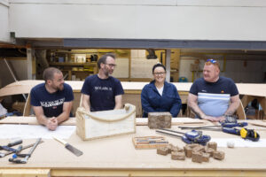 Four adults with light skin smile and engage in conversation while seated at a work bench. The workbench is covered in tools, plans, and blocks of wood.