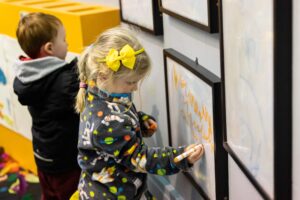 A child with light skin and blonde hair tied into a ponytail uses an orange marker to draw on a wall-mounted board.