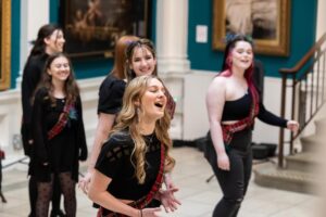 A group of singers wearing black outfits and tartan sashes stand a sing in a gallery.