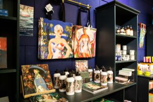 A retail display featuring bags, water bottles, books, and calendars. The merchandise features details of colourful paintings.