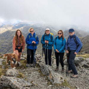 Five adults with hiking gear standing on rocky terrain with a brown dog. Behind them are several mountain peaks and a sky filled with low grey clouds.