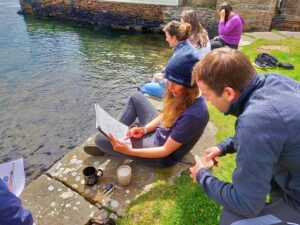 A small group of adults sit on a stone wall overlooking a shallow body of water. Two of the adults discuss some notes written in a notebook.