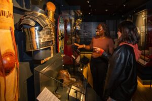 Two young adults - one with dark skin and long brown hair, and another with medium skin and medium-length brown hair - stand and talk while looking at replicas of Roman armour on display in a museum.