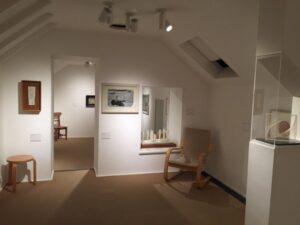 White walls with paintings on them and a sculpture in an alcove next to a wooden chair. White lights hang from the ceiling with bright white spotlights on the artwork.