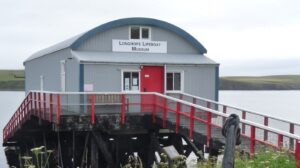 A single-storey building made of grey metal, with a red door and a sign with the text "Longhope Lifeboat Museum". It stands on a small jetty which extends over a body of water.