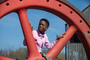 A child with medium-dark skin and a pink t-shirt looks out between the spokes of a large metal wheel.