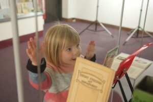 A young, fair skinned child with bobbed blonde hair, presses her hands and face against a glass display cabinet. Her nose is squashed against the glass. Several colourful pamphlets are out of focus in the display cabinet.