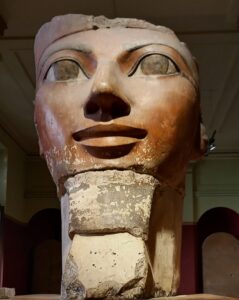 A large sculpture of a face with medium-dark skin and large eyes with eyelids outlined in black. The top of the sculpture is missing and the bottom of the face is worn away. It sits on a worn stone plinth with a sign underneath.