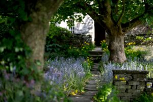Blue and yellow flowers line an uneven stone path which runs between two leafy trees. The path leads to a stone building.