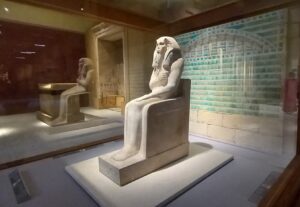A stone carving of an ancient Egyptian king sitting. Behind are pale blue tiles.