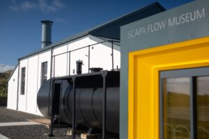 A large black cylindrical boiler situated in front of a single-storey building with white walls and a grey roof. In the foreground is a yellow-trimmed doorway which bears the text "Scapa Flow Museum".