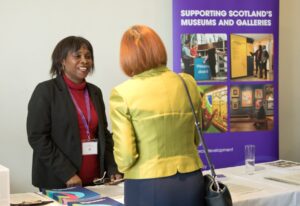 An adult with medium-dark skin and medium-length brown hair stands at a table while smiling at an adult with red hair and a light green jacket. Next to them is a purple banner with the text "Supporting Scotland's Museums and Galleries".