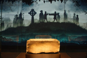 The Stone of Destiny on display at Perth Museum. The large block of stone is displayed in front of a wall with silhouettes of people walking being projected on to it.