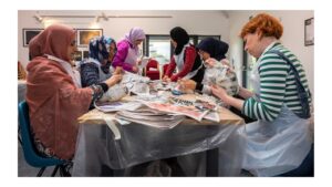 A group of 6 people are sitting at a table creating 3d objects from newspapers. Five people are wearing different coloured headscarves and have medium skin tone. One person has short orange hair and light skin tone.