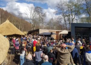 A crowd of people standing together and watching performers on a stage. A replica Iron Age roundhouse and another wooden building can be seen in the background.