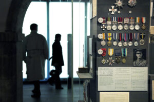 A case displays rows of military medals with colourful ribbons. In the background, behind the case, are the silhouettes of two adults.