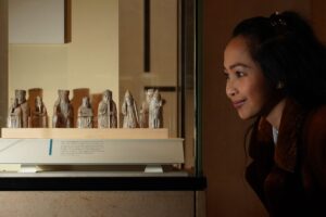 An adult with medium-light skin and black hair tied into a ponytail smiles as they look into a glass case containing ten medieval chess pieces carved from bone.