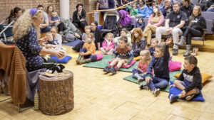 A group of children sit on colourful mats, watching as a seated adult with light skin and long curly grey hair gestures to them with their hands.