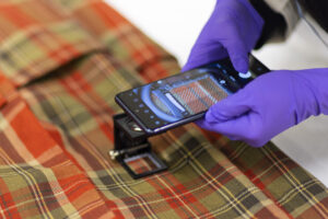A pair of hands in purple nitrile gloves holds a smartphone over a length of red and green tartan fabric.