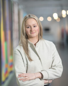 A mid length portrait of a person with long blonde hair, light skin tone, a nose piercing, wearing a white jacket.