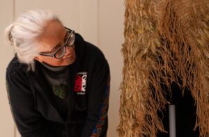 An adult with medium-tone skin, a chin tattoo, and white hair tied into a bun observes a Maori cloak made of brown feathers.