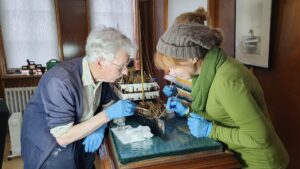 Two adults use cotton buds to clean a model ship. One adult has light skin, short grey hair, and glasses. The other has light skin, red hair in a beige woolen hat, and a green jumper.