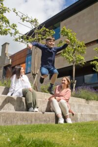 An adult with light skin and tied-back wavy red hair and an adult with light skin and medium length dark brown hair sit on stone steps in a garden. A child with light skin and short brown hair jumps between them.