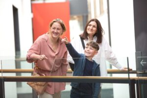 An adult with light skin and tied-back wavy red hair, an adult with light skin and medium length dark brown hair, and a child with light skin and short brown hair stand together on a raised walkway within a spacious gallery. The child is pointing to something high above them.