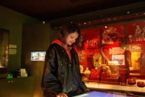 A young adult with medium-light skin tone and shoulder-length brown hair interacting with a screen on a counter in the middle of a museum gallery.