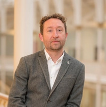 A person with light skin and short curly brown hair, who is wearing a grey suit jacket and white shirt, leans against a railing and stares directly in to the camera.