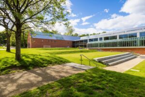 A large L shaped building with red brick walls, a grey metal roof, and long rows of large windows. In front of the building is a sloping lawn.