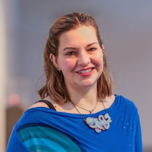 A light skinned person with shoulder length brown hair, partial tied back in a ponytail, wearing a bright blue top with a mandala pattern on it.
