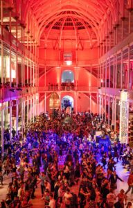 A large three-storey atrium with two floors of balconies supported by narrow white pillars. The atrium is filled with dancing people.