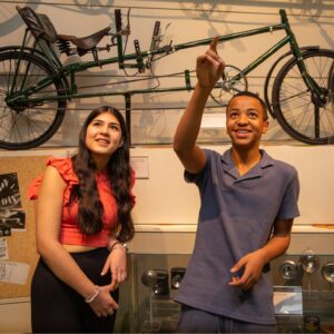 A teenager with medium-light skin and long brown hair and a teenager with medium-dark skin and short brown hair stand in front of a display of bicycles. The teenager with medium-dark skin points up at the ceiling and smiles.