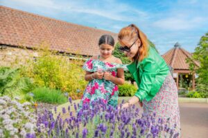 An adult with medium-light skin, long orange hair and a green jacket and a child with medium-light skin, brown tied-back hair and a bright floral dress stand and smile as they play with the flowers of a lavender plant.