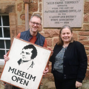 Two adults with light skin and grey hair stand in front of a red stone building. One of the adults hold a sign which features an illustration of 18th century Scottish poet Robert Burns and the text "Museum Open".