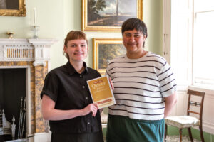 Two adults stand together in an ornately decorated room which has a fireplace and gold-framed paintings hanging from the wall. One adult has light skin, straight light-brown tied-up hair, and is holding up a booklet titled "How to Queer your Historic House". The other adult has medium-tone skin and short, straight, dark grey hair.