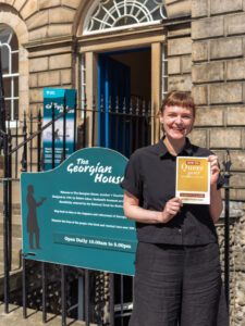 An adult with light skin, straight light-brown tied-up hair, and a black shirt smiles while holding up a yellow booklet with the title "How to Queer your Historic House." They are standing in front of a stone building with ornate iron rails, a large open door, and a sign reading "The Georgian House".
