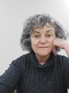 An adult with light skin tone and short grey hair. They are wearing a grey turtle neck jumper.