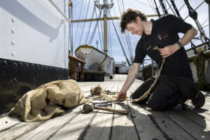 An adult with light skin and short brown hair kneels on the wooden deck of a ship with a length of rope, a hammer, and metal pegs.