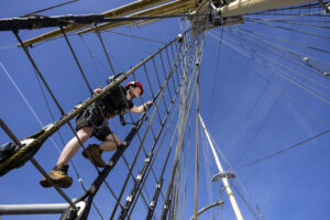 An adult with light skin and a red hard hat climbs up the rigging of a ship.