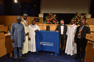 6 people stand in an auditorium around a Benin Bronze sculpture placed upon a plinth covered in a blue table cloth that has "University of Aberdeen" written on it. 5 of the people have dark skin, 1 has light skin. The people are all wearing examples of formal dress in either the western european style or North West African style.