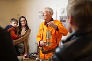 An adult with light skin, grey hair, an orange jumpsuit, and a white hard hat shows a metal safety lamp to a group of adults and children.