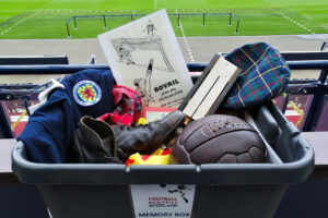 A black plastic tub containing objects associated with Scottish football in the mid-20th century. This includes a leather football, a football boot, a cap, and a wooden rattle.