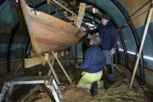 Two adults constructing a wooden rowing boat propped up on wooden stilts.