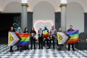 A group of nine smiling adults stand in a line and hold Pride Progress flags.