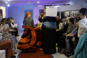 A young person with medium skin tone is walking down a runway wearing a red head scarf and a flowing red and black dress. An adult is standing next to them and holding a sign that reads: "Article 27. Everyone has the right to freely participate in the cultural life of the community, to enjoy the arts and share in scientific advancement and all its benefits."