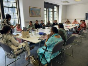 A group of adults with medium skin tone is gathered around a large table. They are having conversations across the table.