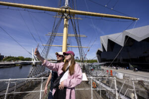 Two adults wearing sunglasses and baseball caps - one with light skin and a brown beard, and one with light skin and long straight brown hair - pose for a selfie on the deck on a sailing ship.