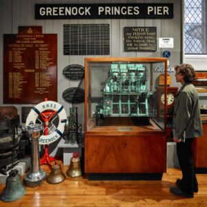An adult with light skin and wavy shoulder-length brown hair stands next to a display of maritime objects. The display includes ship bells, a life rings, and a large sign with the text "Greenock Princes Pier".